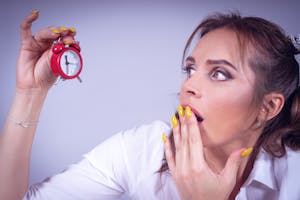 FOMO A surprised woman with yellow nails holds a small red alarm clock against a white backdrop.