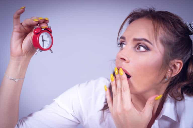 FOMO A surprised woman with yellow nails holds a small red alarm clock against a white backdrop.